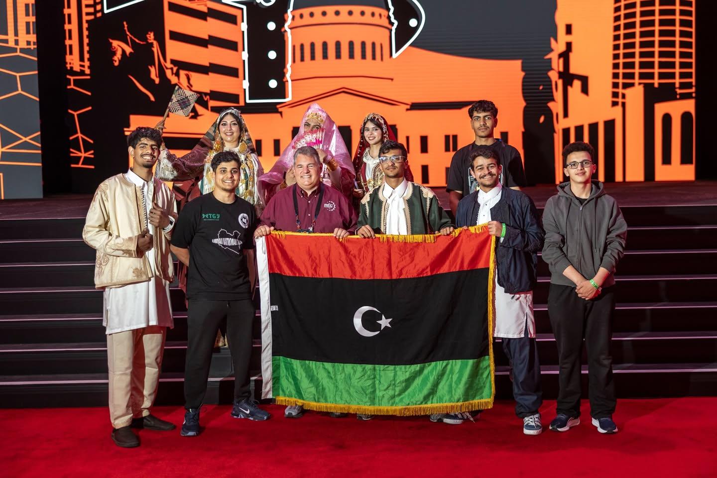أعضاء من الفريق الليبي يحملون علم ليبيا أمام المسرح في بطولة العالم VEX للروبوتات، وخلفهم ثلاث نساء بالزي التقليدي، سانت لويس، أبريل 2026